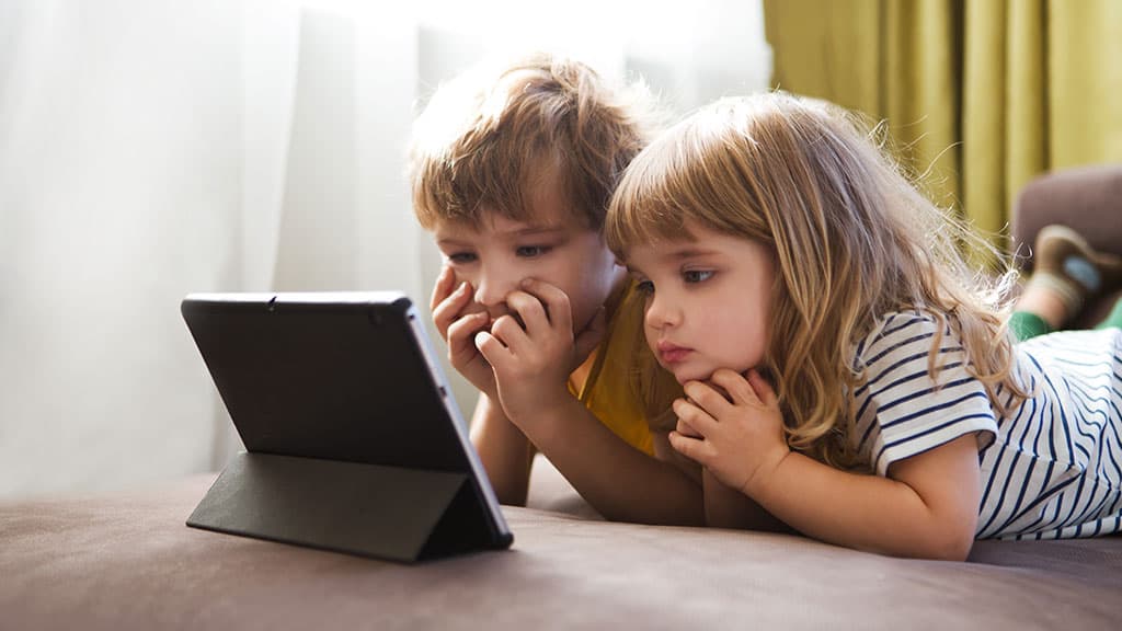 dwoje małych dzieci leży na kanapie i patrzy z bliska w ekran tabletu